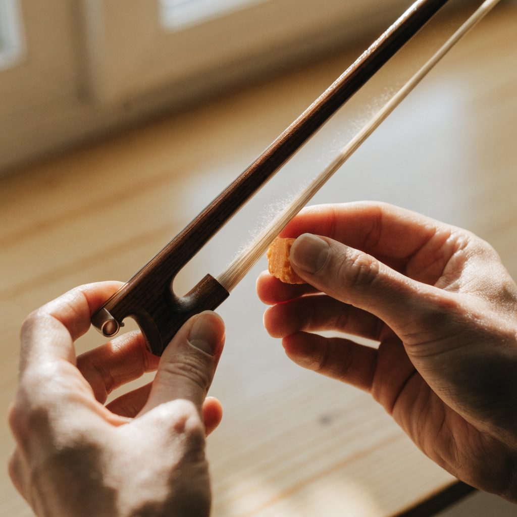 Close view of adult hands drawing a cake of rosin along the hair of a violin bow on a wooden workbench.