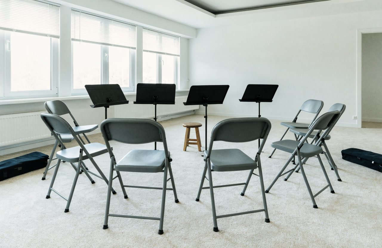 A semicircle of empty folding chairs and music stands in a bright open room, with two soft instrument cases resting on the floor nearby.