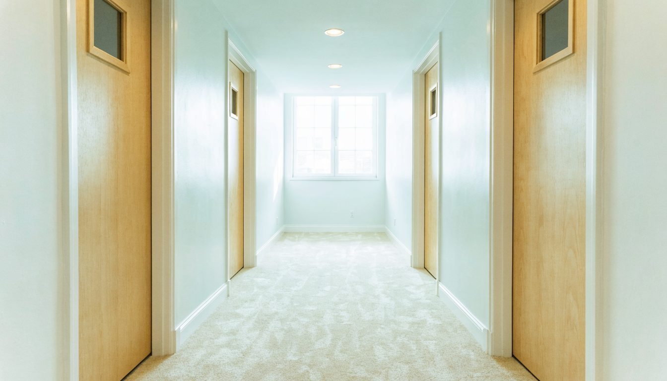 A bright corridor inside the school with five plain teaching room doors along it, pale carpet, white walls, and daylight coming from an open door at the far end.