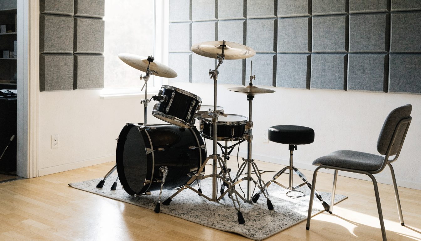 A drum kit with three cymbals set up on a rug in a small room, its upper walls covered in grey acoustic foam panels, with a drum stool and a chair beside it and daylight coming through a window at the left.