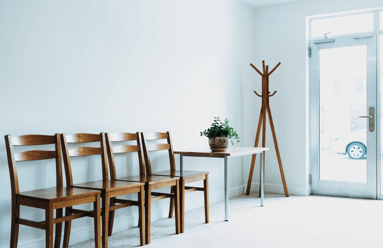 The small front waiting area of the school, with four wooden chairs against a pale wall, a low table holding a plant, a coat stand, and a glass entrance door letting in bright daylight.
