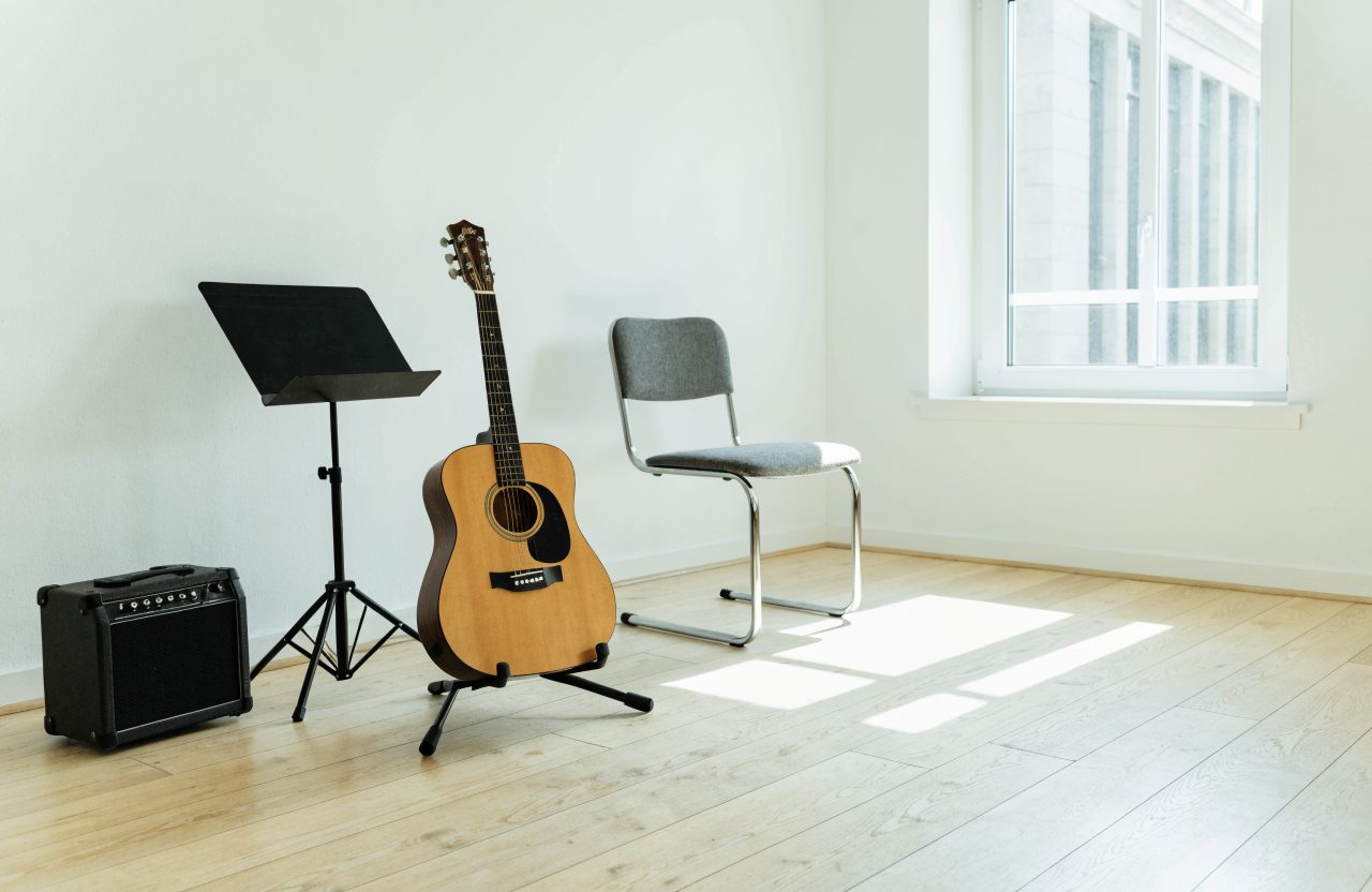 A small bright teaching room with an acoustic guitar resting on a floor stand, a folding chair, a small amplifier on the floor and a music stand beside a window.