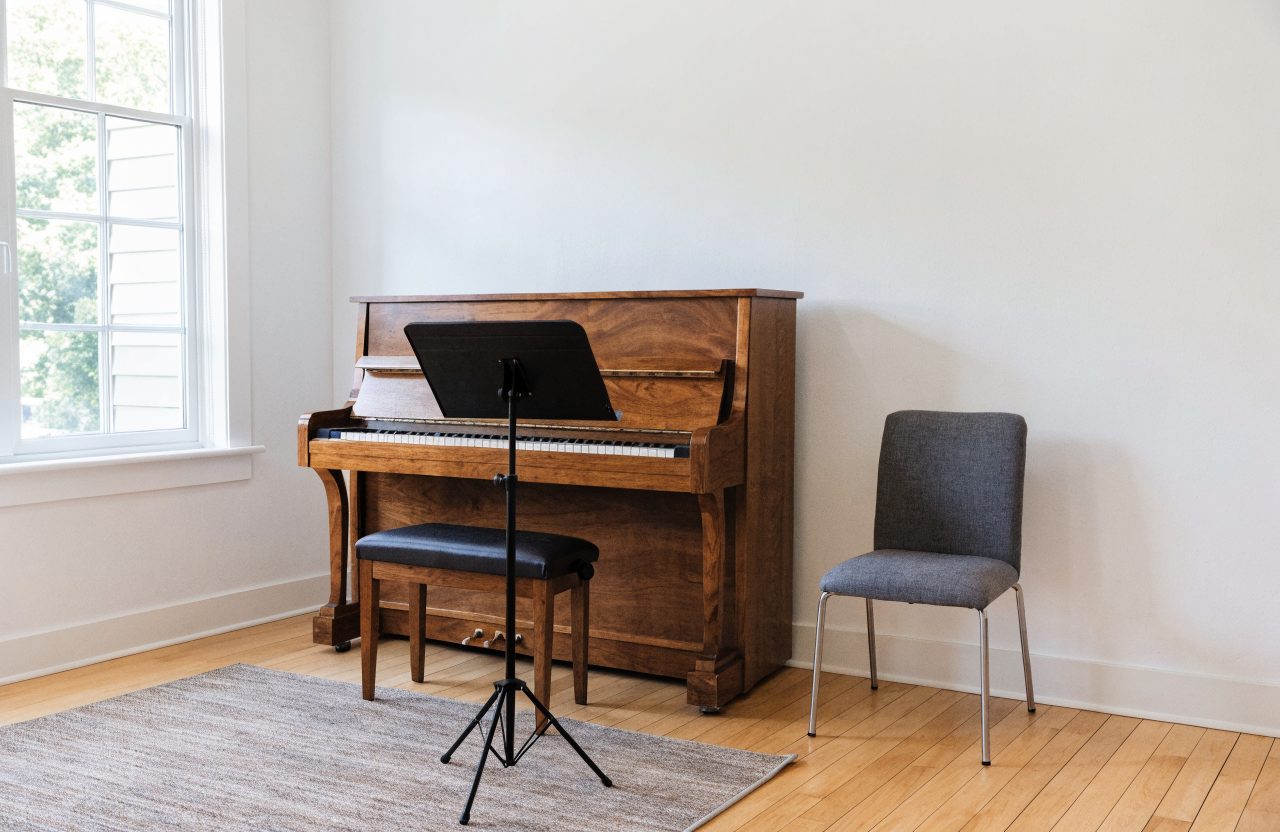A small private lesson room with an upright piano against a white wall, a padded bench, a metal music stand and one folding chair, lit by daylight from a window at the left.